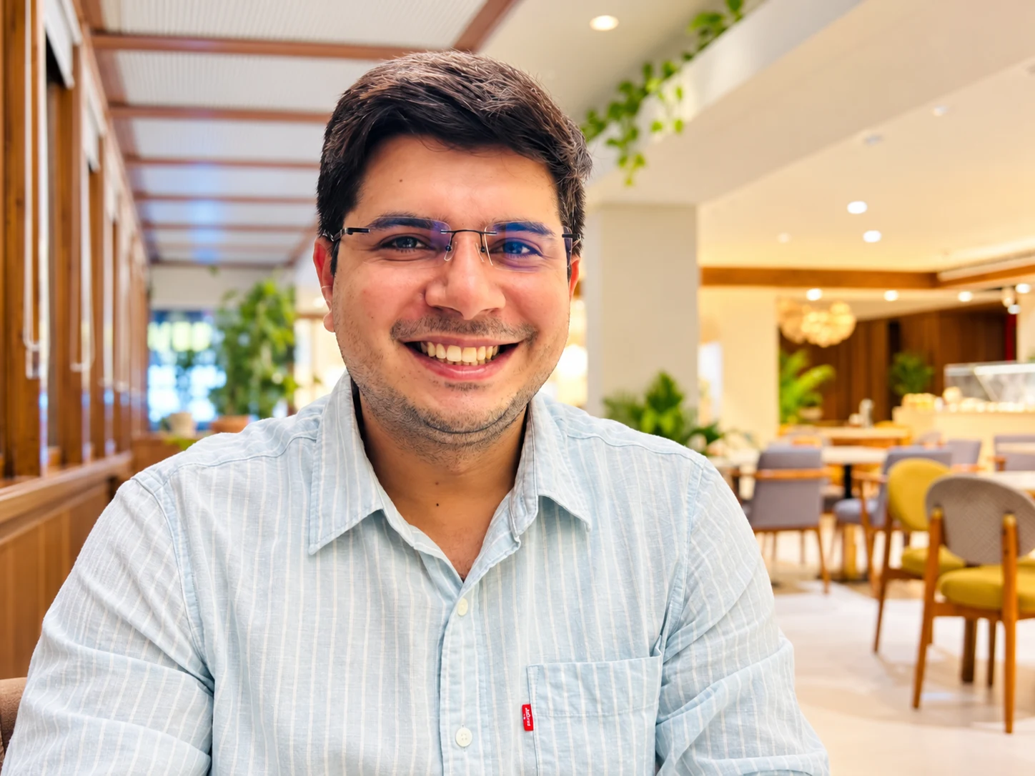 Sanu Siddharth smiling at a café, wearing glasses and a light striped shirt